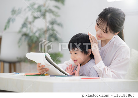 A toddler enjoys learning while being watched by his mother (mother) and opens his notebook. Praise the child. 135195965