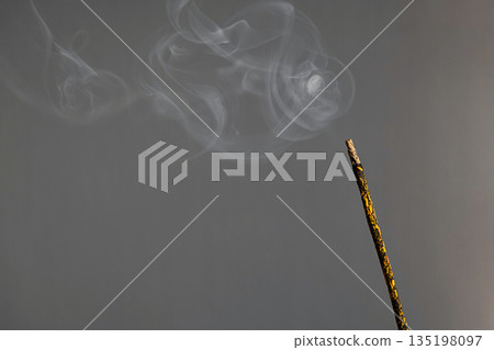 Lit incense stick releasing delicate smoke against calm gray background. 135198097
