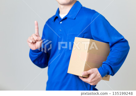 A man in the logistics and transportation industry holding a cardboard box and pointing his index finger up 135199248