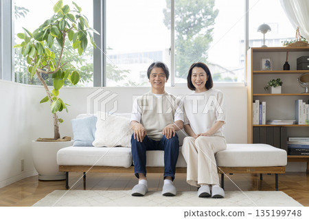 一對五十多歲的夫婦並排坐在沙發上，對著鏡頭微笑。 135199748