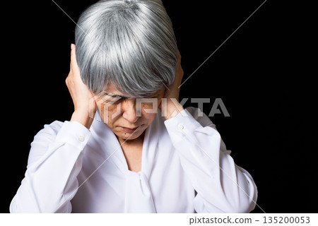 A gray-haired senior woman covering her ears with both hands and frowning A gray-haired senior woman covering her ears with both hands and frowning 135200053