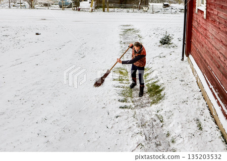 Sweeping snow from concrete walkway toward entrance door of rural house man uses broom he is lightly but warmly dressed. 135203532