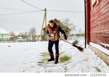 Homeowner clears snow from garden path using broom wearing warm vest and winter boots. Homeowner clears snow from garden path using broom wearing warm vest and winter boots. 135203535