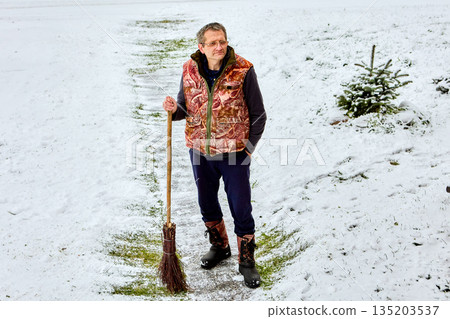 Man standing on snowy path outdoors holding broom in hand near small evergreen tree on side of walkway in winter landscape. Man standing on snowy path outdoors holding broom in hand near small evergreen tree on side of walkway in winter landscape. 135203537