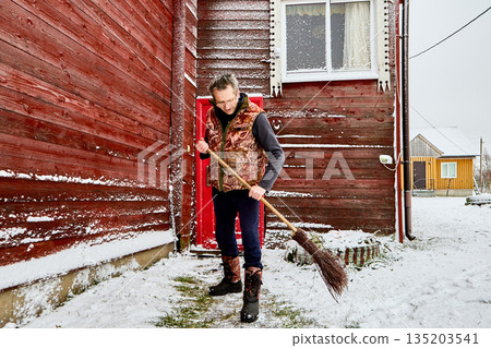 Homeowner sweeps snow from entrance path with birch broom, standing in front of rural house clad with painted red boards, wearing warm vest and winter boots. 135203541