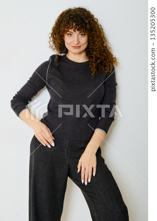 Tricky woman in dark sweater and matching trousers stands gracefully against white wall Tricky woman in dark sweater and matching trousers stands gracefully against white wall 135205300