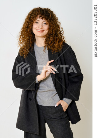 Woman in a stylish black coat and grey outfit poses confidently against a plain background 135205301