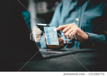 Person Using Smartphone with Digital Checklist Overlay Near Laptop Person Using Smartphone with Digital Checklist Overlay Near Laptop 135206733