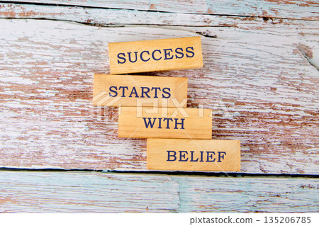 Wooden blocks are stacked vertically, each inscribed with a word. Together, they form the message SUCCESS STARTS WITH BELIEF on an aged, painted surface, evoking tenacity 135206785