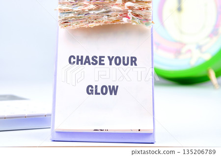 A desk calendar displays the message Chase Your Glow. Multicolored scraps sit on top. A desk and clock appear in the background, bathed in gentle light A desk calendar displays the message Chase Your Glow. Multicolored scraps sit on top. A desk and clock appear in the background, bathed in gentle light 135206789