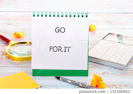A vibrant notepad boldly states GO FOR IT while a magnifying glass, calculator, pencil and paper scraps are on a bleached wood desk, inspiring success and motivation 135206802