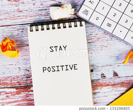 A spiral notebook with the inspiring words STAY POSITIVE is on a weathered wooden table, next to a calculator and crumpled paper, creating a serene workspace 135206805