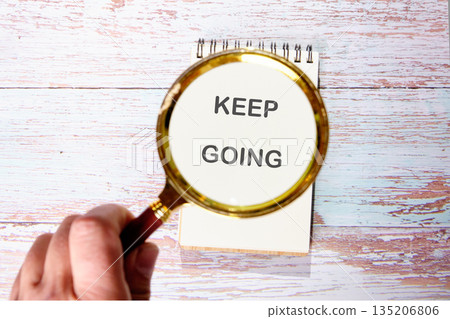 A hand holds a magnifying glass over a notepad. The notepad reads KEEP GOING. The background is a faded wooden surface. It encourages continuous effort and perseverance 135206806