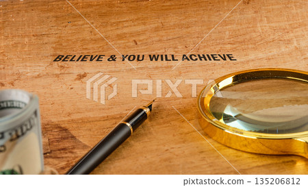 The words BELIEVE and YOU WILL ACHIEVE are displayed, complemented by a golden magnifying glass, a distinguished fountain pen, and a roll of United States currency The words BELIEVE and YOU WILL ACHIEVE are displayed, complemented by a golden magnifying glass, a distinguished fountain pen, and a roll of United States currency 135206812