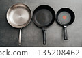 Trio of cookware with matching handles displayed together, Series of three stainless steel frying pans with distinct nonstick and speckled finishes arranged side by side 135208462