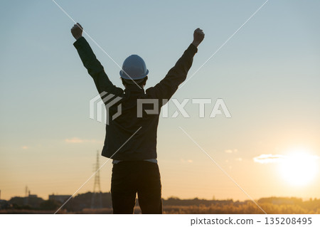 A man in work clothes cheering with the sunset in the background 135208495