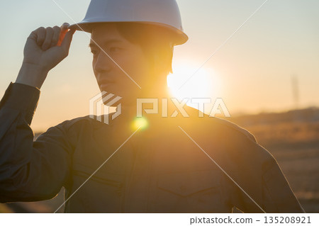 一位身穿工作服的男子站在夕陽下，背景是落日。 135208921