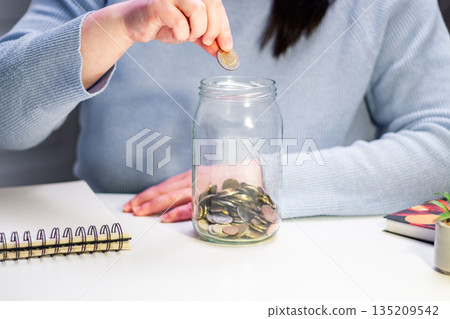 Close up of coins being added to transparent money jar. Represents traditional saving methods and conscious money management for future security 135209542
