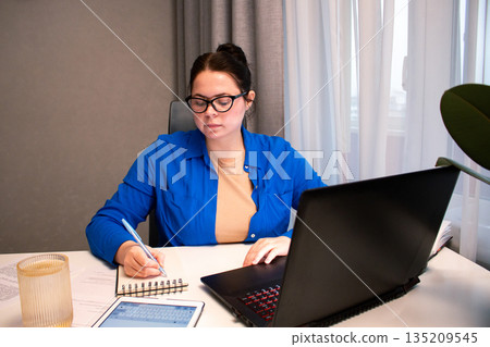 Student attending virtual university class via video conference. Young woman actively participates in online lecture, taking notes on laptop. Distance learning and educational technology concepts 135209545