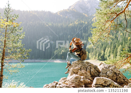 Back view young woman with backpack looking the view of the mountain lake in sunny weather. 135211198