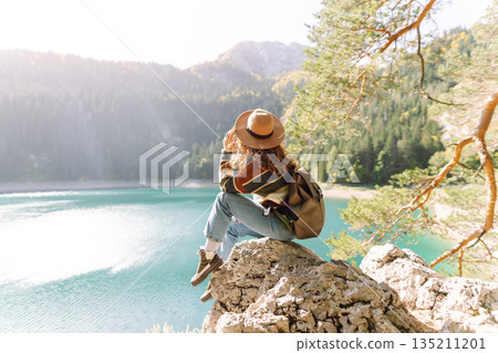 Back view young woman with backpack looking the view of the mountain lake in sunny weather. 135211201