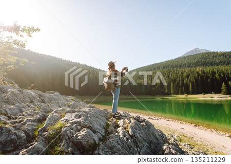 Young woman takes a photos the nice view by the serene lake surrounded by forested mountains Young woman takes a photos the nice view by the serene lake surrounded by forested mountains 135211229