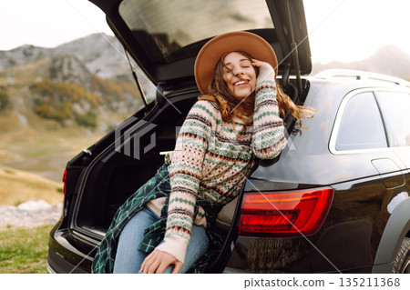 A traveler woman with curly hair and a hat smiles as she relaxes at the back of her black SUV A traveler woman with curly hair and a hat smiles as she relaxes at the back of her black SUV 135211368