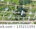 Blue glass wind chimes hanging from a wooden frame, green summer scenery 135211393