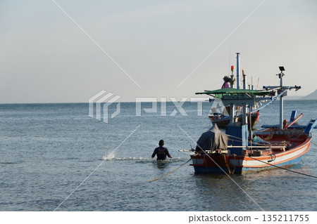 The fisherman and the sea.A man is fishing with a net in the sea, next to a colorful wooden fishing boat. 135211755