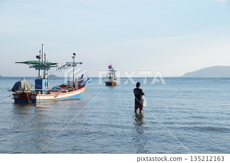 The fisherman and the sea.A man is fishing with a net in the sea, next to two colorful wooden fishing boats. The fisherman and the sea.A man is fishing with a net in the sea, next to two colorful wooden fishing boats. 135212163