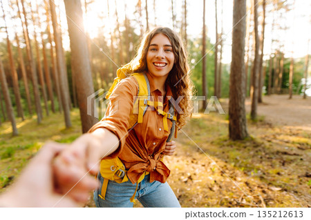 Follow me! Young woman joyfully exploring a sunny forest trail with a backpack, holding hands 135212613