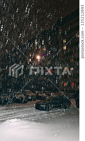 a view of a nighttime city courtyard with a car park during a winter blizzard and snowfall. 135212693