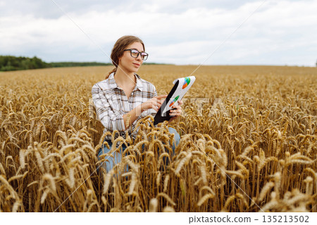 Young female farmer checking crop growth with clipboard in golden wheat field. Smart farming. 135213502