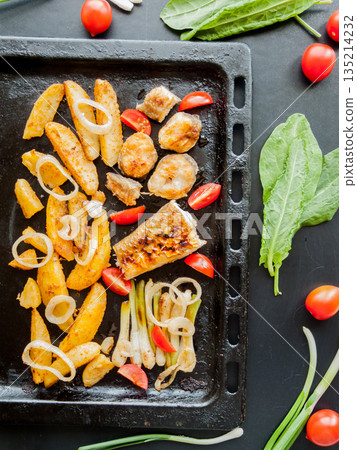Oven pan with sliced baked potatoes, fish steaks and vegetables with onion on table. 135214232
