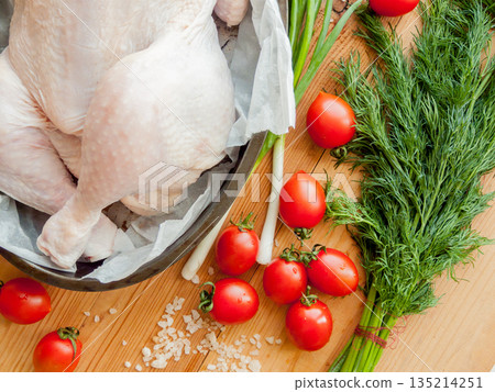 Whole raw chicken laying in the pan with parchment on the wooden table preparing for baking, surrounded by fresh vegetables. Whole raw chicken laying in the pan with parchment on the wooden table preparing for baking, surrounded by fresh vegetables. 135214251
