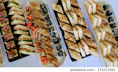 Gourmet catering featuring elegant platters with colorful canapes, triangular sandwiches, and delicate rolled crepes for professional event dining Gourmet catering featuring elegant platters with colorful canapes, triangular sandwiches, and delicate rolled crepes for professional event dining 135215083