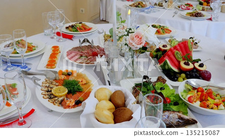 Beautifully decorated table is set for an event in a restaurant with various dishes of meat, fish, salads, fruits, vegetables, bread and flowers 135215087