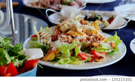 Grilled chicken caesar salad featuring crisp romaine, golden croutons, parmesan shavings, ripe tomatoes on elegant wooden surface 135215101
