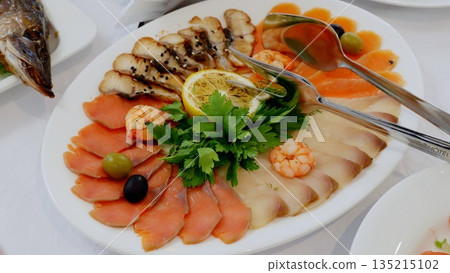 Luxurious seafood platter displaying smoked fish, salmon slices, shrimp, and garnished with lemon and olives on crisp white linen surface 135215102
