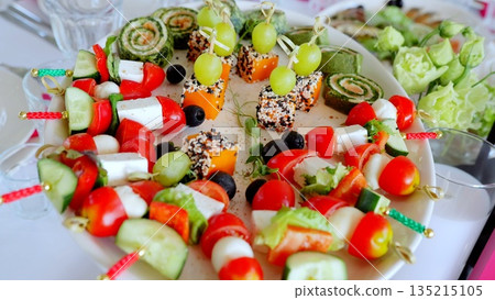 Colorful canapes featuring cheese, tomatoes, cucumbers, olives, grapes, spinach rolls, and pumpkin pieces with sesame seeds arranged on a white plate for festive celebrations 135215105