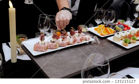 Professional chef wearing white gloves carefully positioning cherry tomato atop canape, completing gourmet appetizer platter for high end catered event 135215127