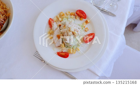 Crisp romaine lettuce tossed with grilled chicken, golden croutons, shaved parmesan, ripe cherry tomatoes resting on pristine white linen, embodying classic caesar salad elegance 135215168