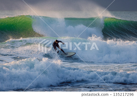 Surfer in wetsuit rides on wave in cold sea. Winter surfing with evening sunlight 135217296