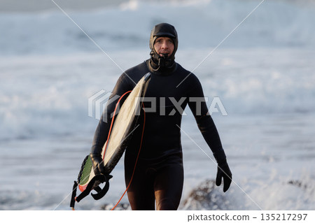 Surfer man with surfboard in winter wetsuit walk on beach Surfer man with surfboard in winter wetsuit walk on beach 135217297