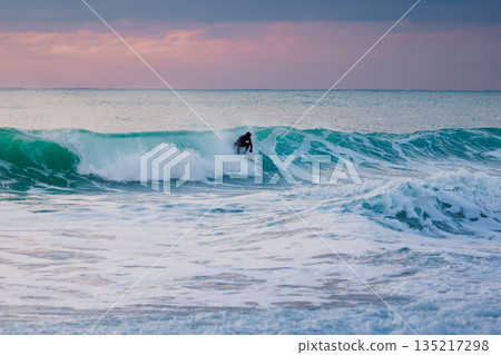 Surfer in wetsuit rides the waves in ocean with sunset tones Surfer in wetsuit rides the waves in ocean with sunset tones 135217298