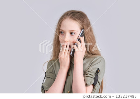 Surprised caucasian young girl talking on smartphone in green shirt on light Surprised caucasian young girl talking on smartphone in green shirt on light 135217483