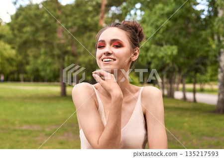 Young caucasian female with red eye makeup smiling in park under bright Young caucasian female with red eye makeup smiling in park under bright 135217593