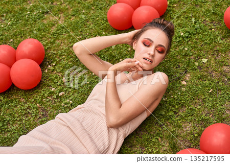 Young caucasian female relaxing on grass with red balloons, eyes closed 135217595