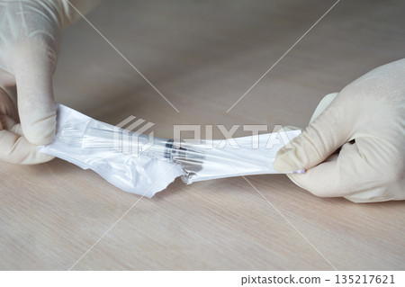 Hands wearing gloves unwrapping sterile medical syringe in protective packaging Hands wearing gloves unwrapping sterile medical syringe in protective packaging 135217621