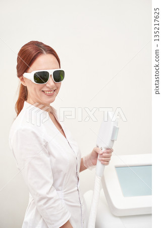 Female caucasian adult in white coat holding cosmetic laser equipment, smiling Female caucasian adult in white coat holding cosmetic laser equipment, smiling 135217625
